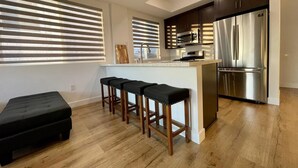 A modern kitchen with a stainless steel refrigerator, a microwave, and a sink. There are three bar stools with cushioned seats.