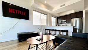 A modern living room with a large flat-screen TV mounted on the wall, a black leather sofa, a round coffee table, and a kitchen area with stainless steel appliances.