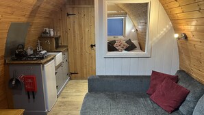 Habitación acogedora y compacta con kitchenette, un sofá gris con un cojín rojo y una pequeña ventana con vistas a un dormitorio.