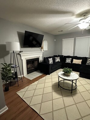 A living room with a fireplace, black sofa, white rug, and a ceiling fan. - Modern house in North Charleston (Charleston)