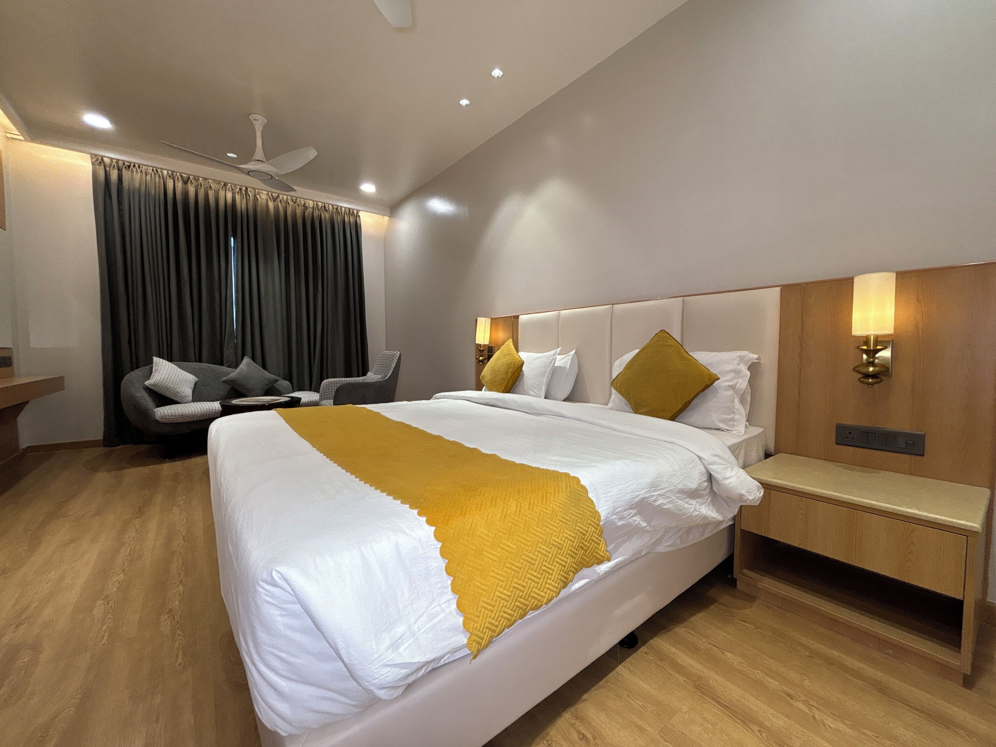 A modern hotel room with a large bed, a bedside table, a chair, a desk, a lamp, and a ceiling fan.