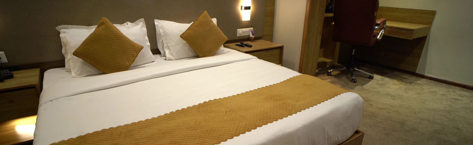 Uma cama bem arrumada com roupa de cama branca e cor de mostarda, uma mesinha de cabeceira com um controle remoto e uma cômoda de madeira.