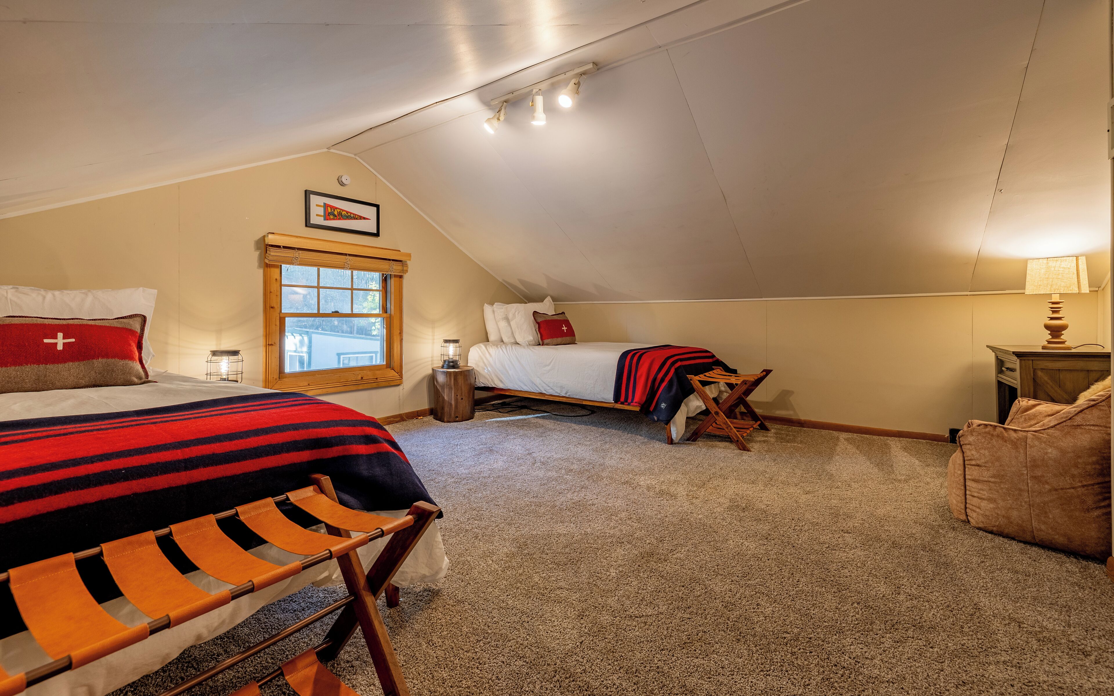 Een gezellig slaapkamer op zolder met een schuine kap, een groot raam, een bed met een gestreept dekbed, een houten stoel en een lamp.