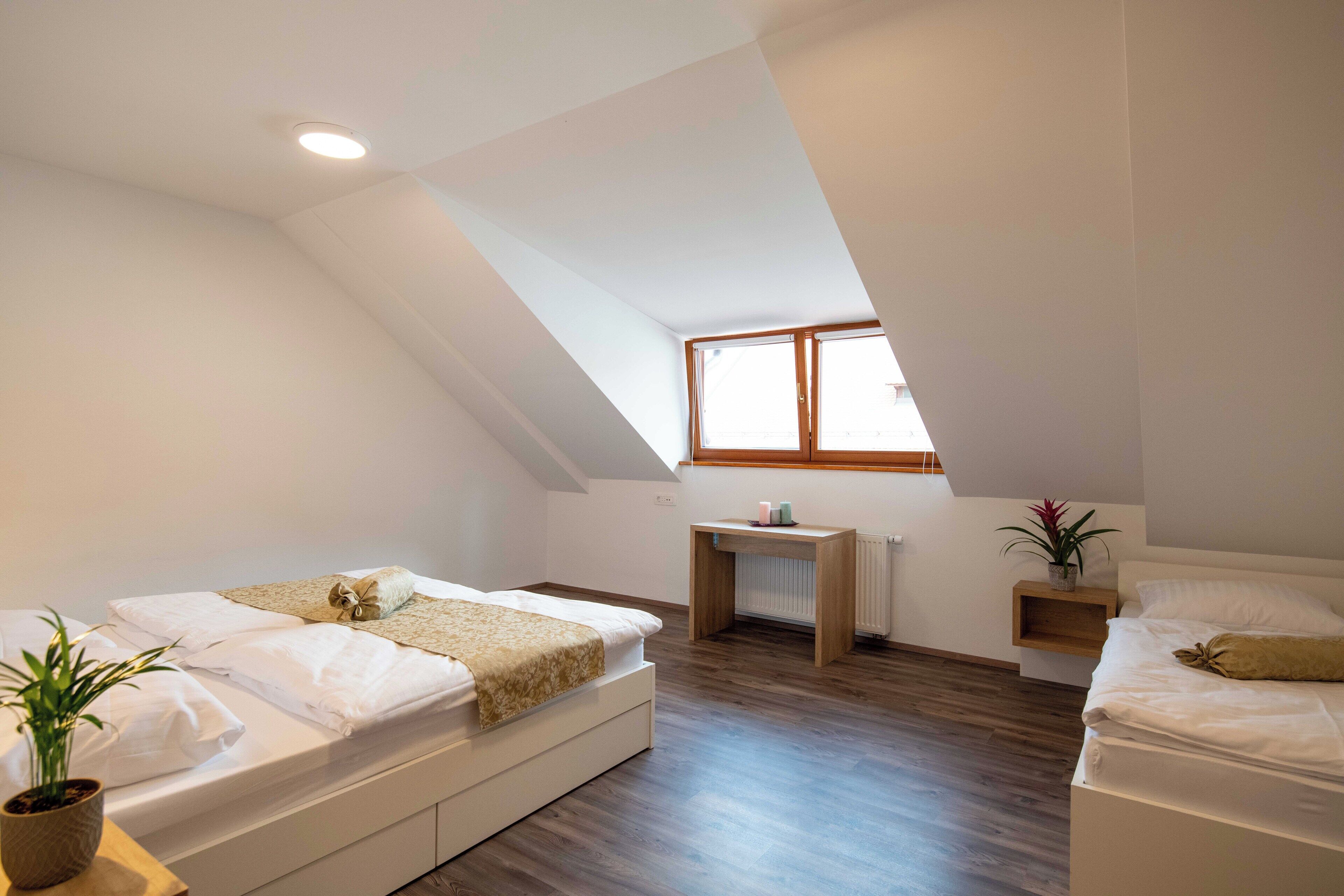 Un dormitorio ático moderno y bien iluminado con dos camas, suelo de madera, una mesita y una ventana que deja entrar luz natural.