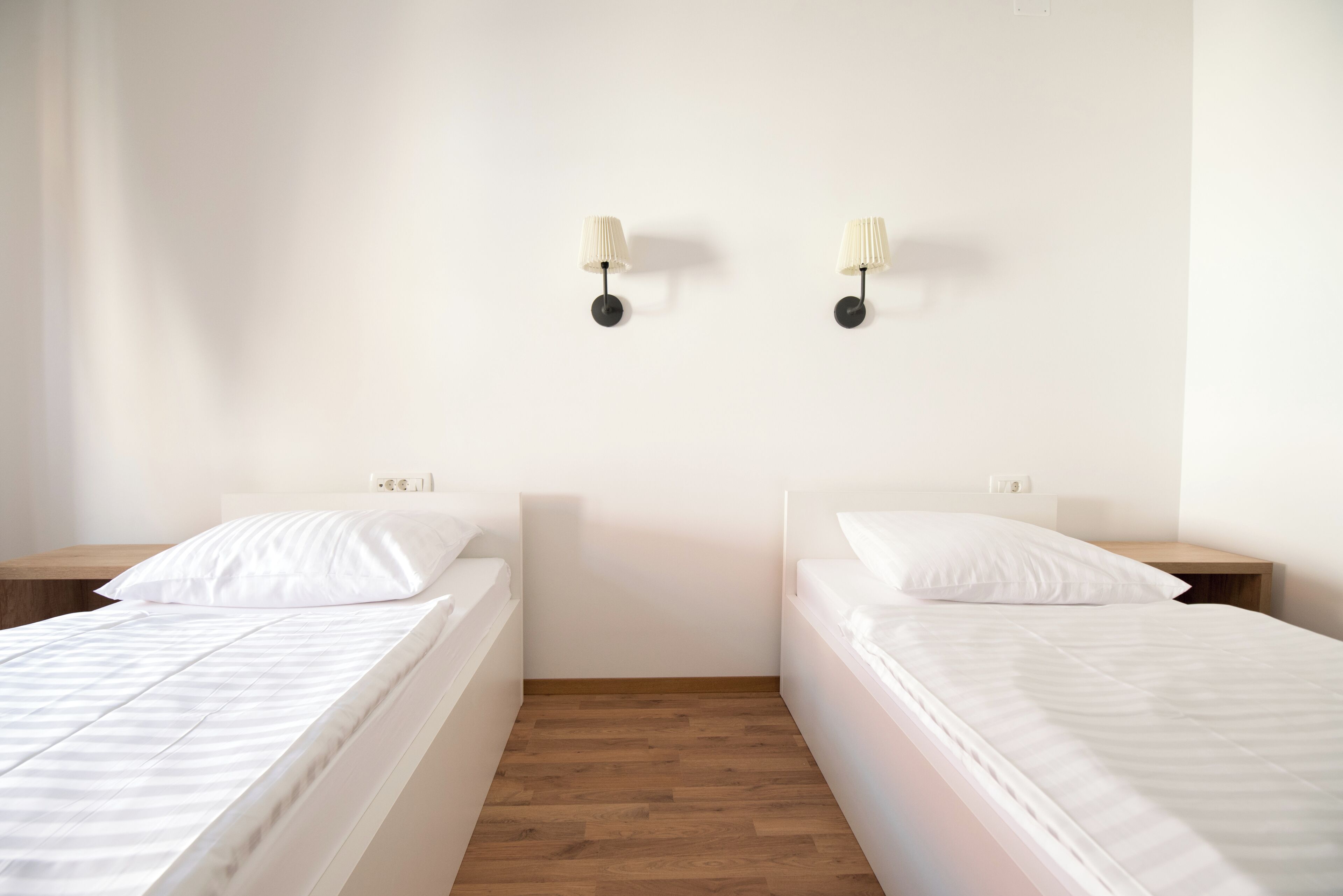 A bedroom with two single beds, wooden flooring, white walls, and two wall-mounted lamps.