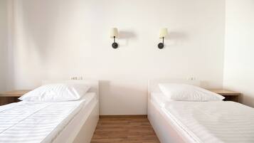A bedroom with two single beds, wooden flooring, white walls, and two wall-mounted lamps.