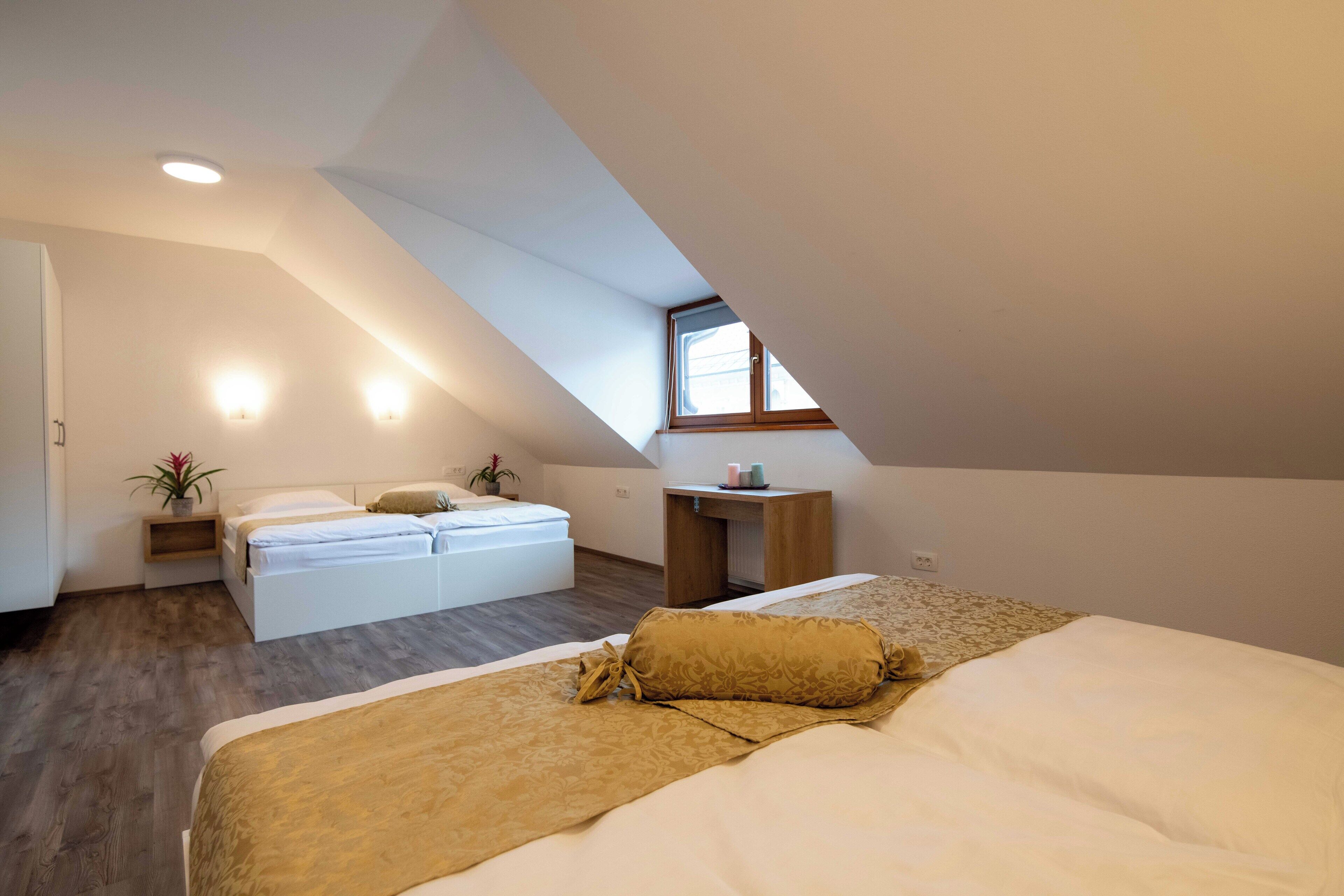A bedroom with a sloped ceiling, a bed with a beige and white duvet, a bedside table with a lamp, and a small wooden desk with a chair.