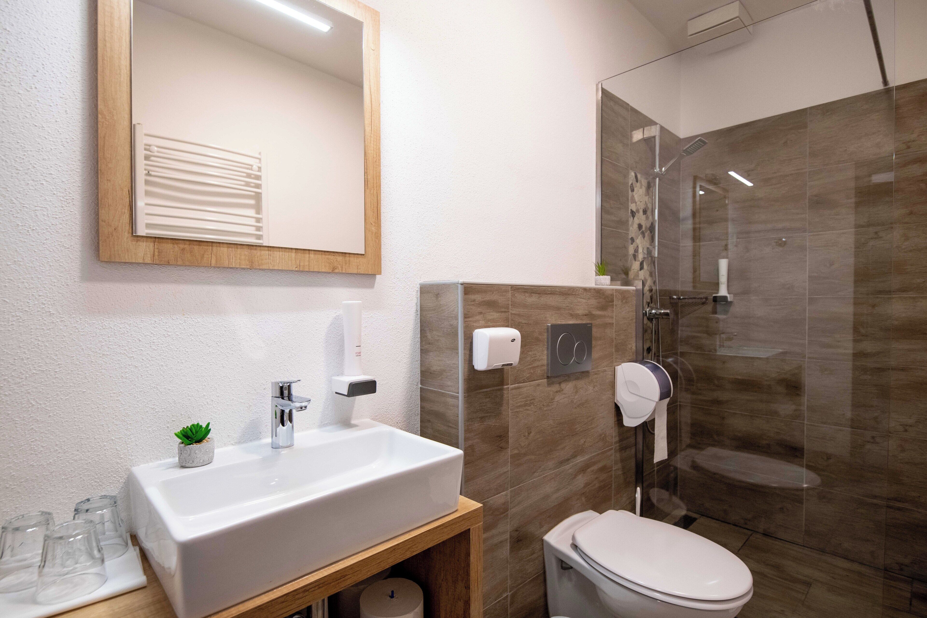 Un baño moderno con un espejo grande, un lavamanos blanco, un inodoro y una ducha con mampara de vidrio.