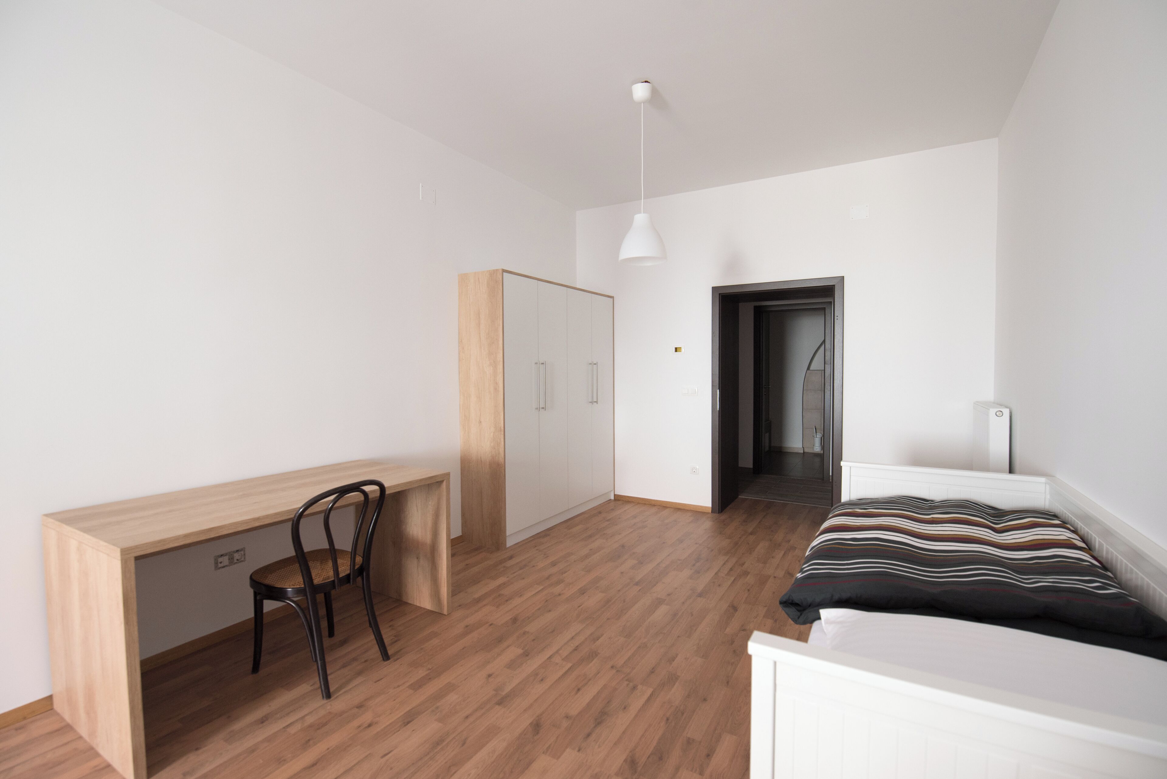 A bedroom with a wooden desk, a chair, a bed with a striped pillow, and a wardrobe.