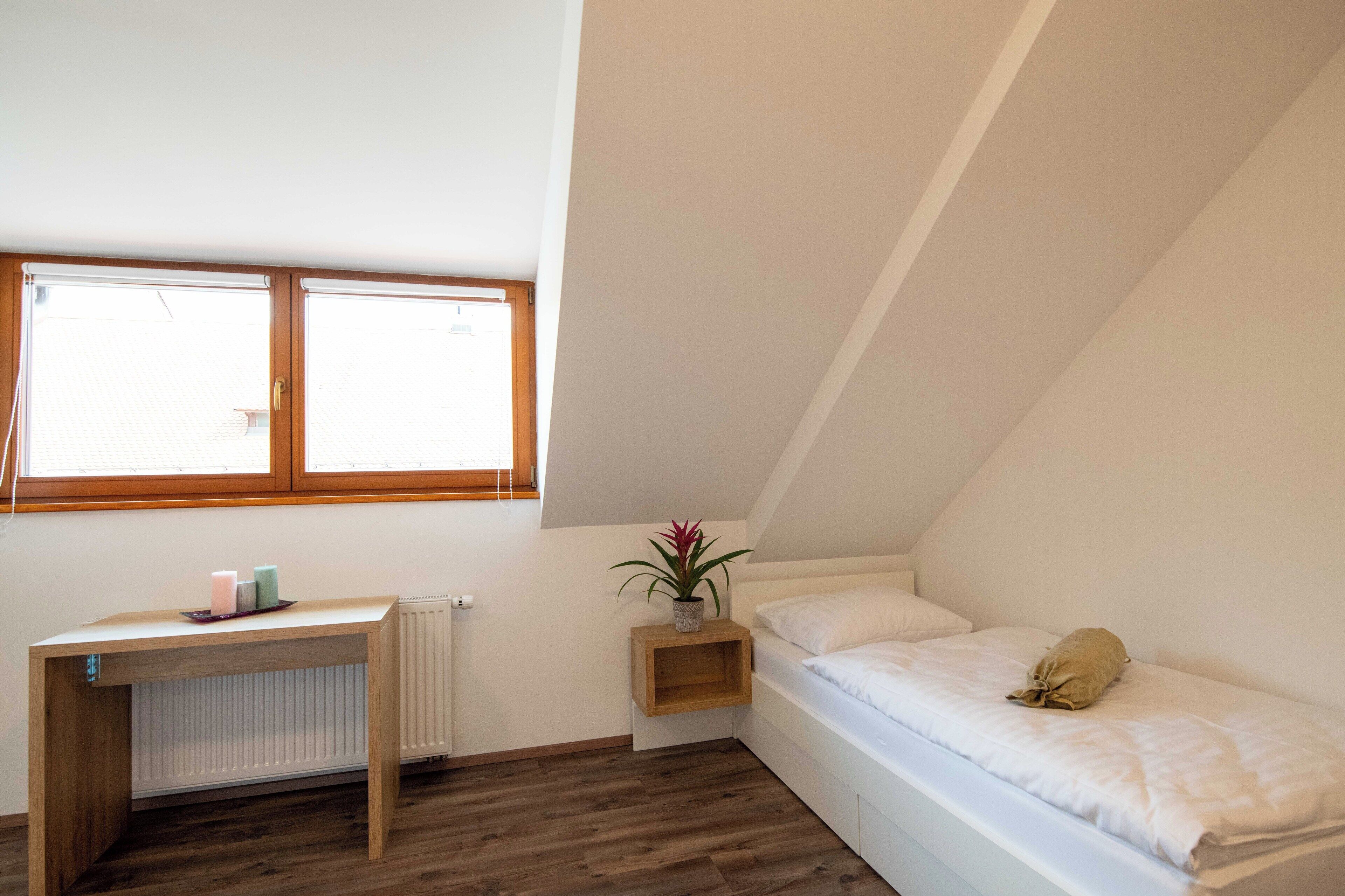 Ein kleiner, gut beleuchteter Raum mit schrägem Dach, einem Einzelbett, einem Nachttisch mit Pflanze und einem Fenster, das für natürliches Licht sorgt.