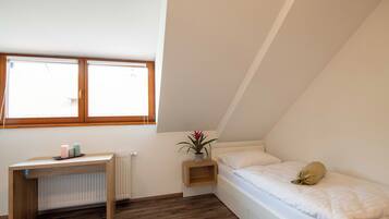 Ein kleiner, gut beleuchteter Raum mit schrägem Dach, einem Einzelbett, einem Nachttisch mit Pflanze und einem Fenster, das für natürliches Licht sorgt.