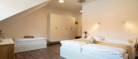A modern bedroom with a large bed, a smaller bed, and a desk. There is a wardrobe and a plant in the room.