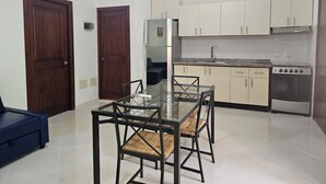 Cocina compacta con armarios blancos, estufa a gas y refrigerador. Hay una mesa de vidrio con dos sillas.