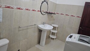 Un baño con lavamanos blanco, un espejo, toallero, una pequeña estantería y dispensador de toallas de papel.
