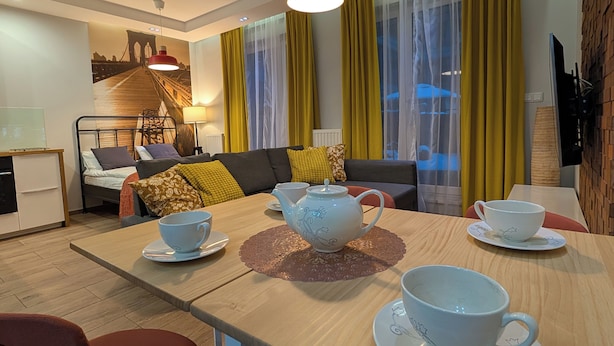 Una sala de estar moderna con sofá, una mesa de comedor con teteras y tazas, cortinas amarillas y un mural con vista a la ciudad.
