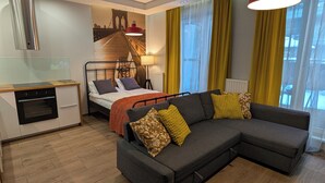 Habitación de hotel moderna con cama, sofá, zona de cocina y un gran ventanal con cortinas.