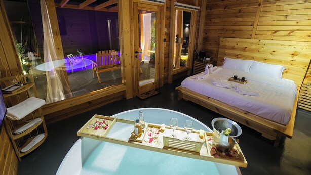 A wooden cabin with a bed, a dining table with a tray set, and a large window. - elmurooj cottage (Jeddah)