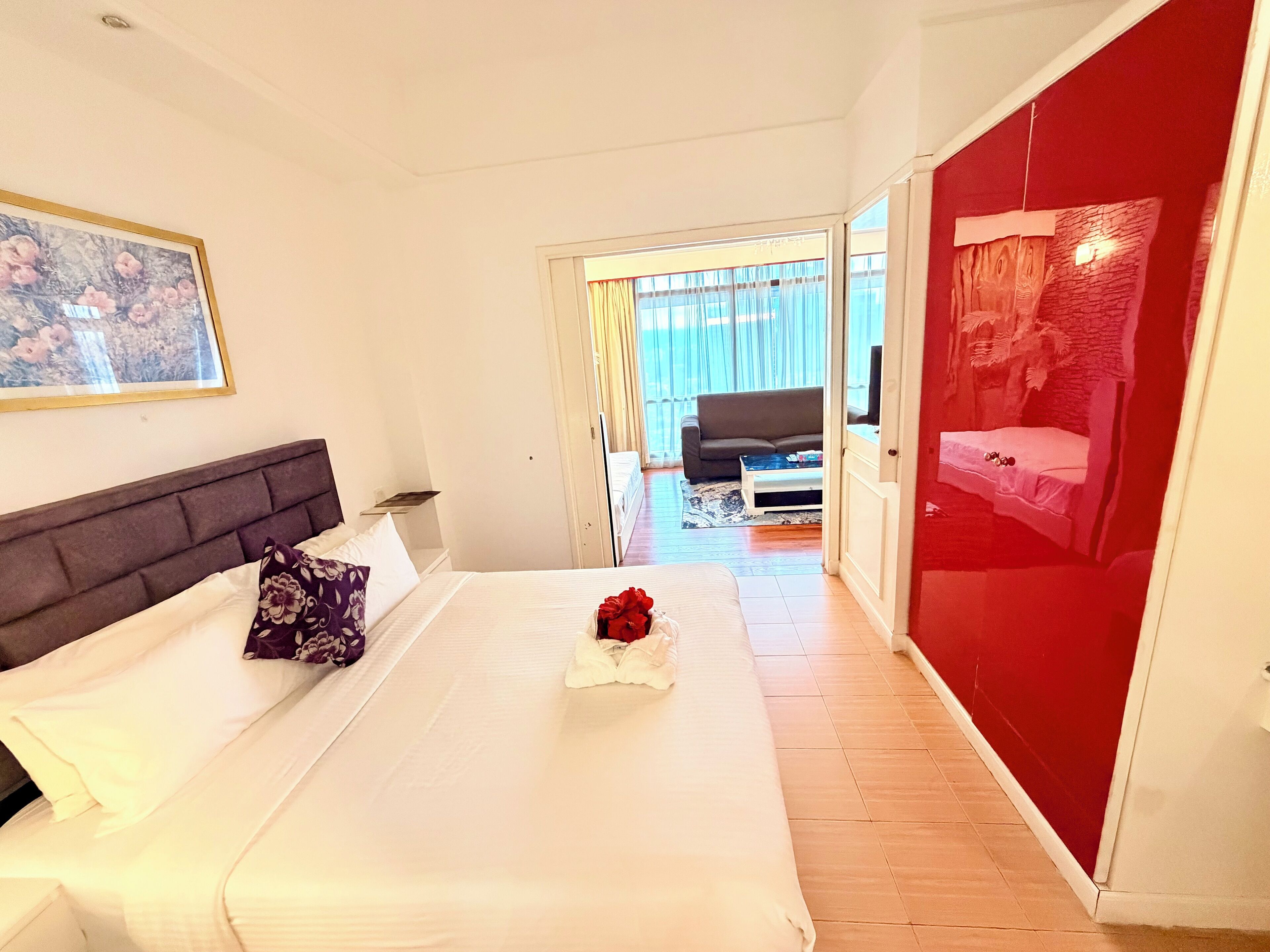 A hotel room with a bed, a nightstand, a painting, a red wardrobe, and a view of the living area.