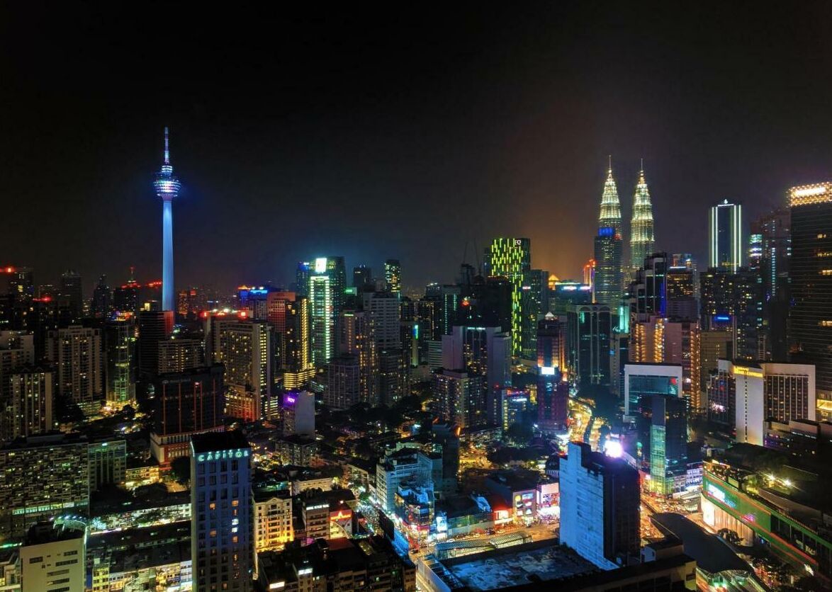 A cityscape at night with illuminated buildings, including a prominent tower and two distinctive twin towers.