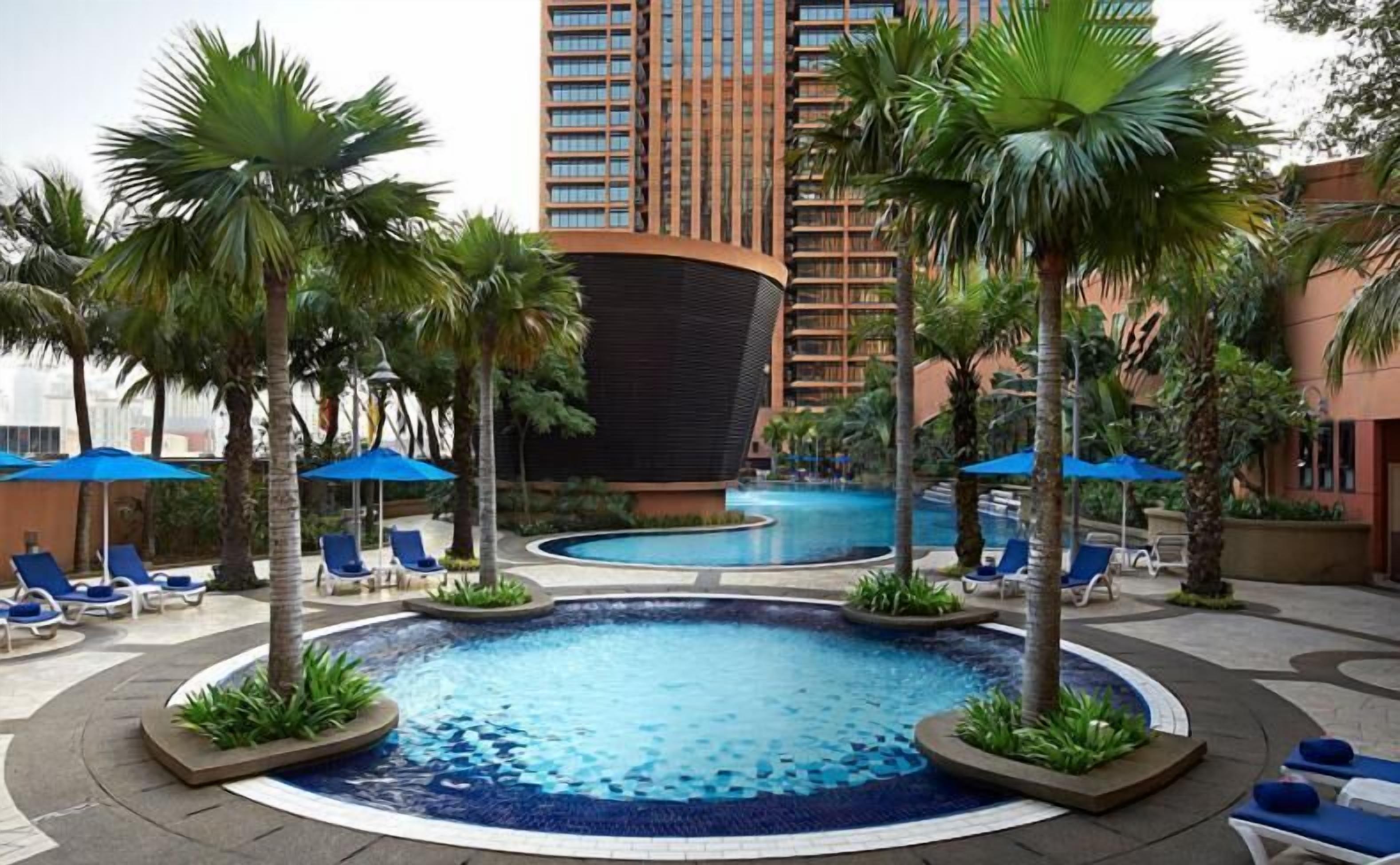 Ein Hotelpoolbereich mit Liegestühlen und Sonnenschirmen, umgeben von Palmen und einem modernen Gebäude im Hintergrund.