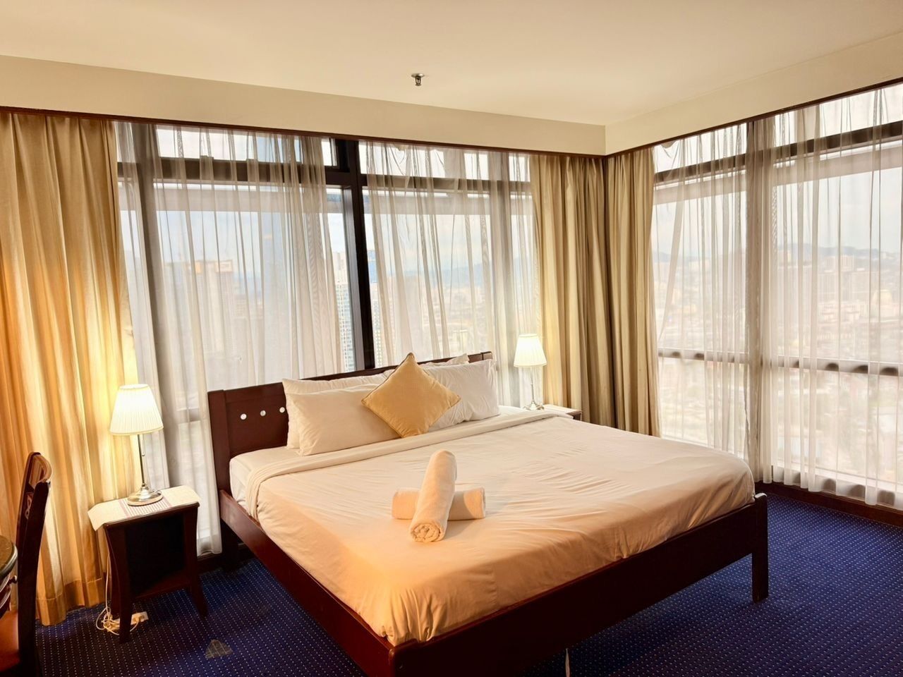 Une chambre d’hôtel avec un grand lit, une table de chevet avec une lampe et une vue sur la ville à travers de fines rideaux.
