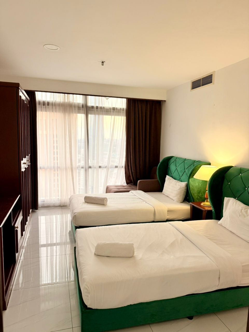 Habitación de hotel con dos camas, un armario, una ventana con cortinas y una lámpara en la mesita de noche.