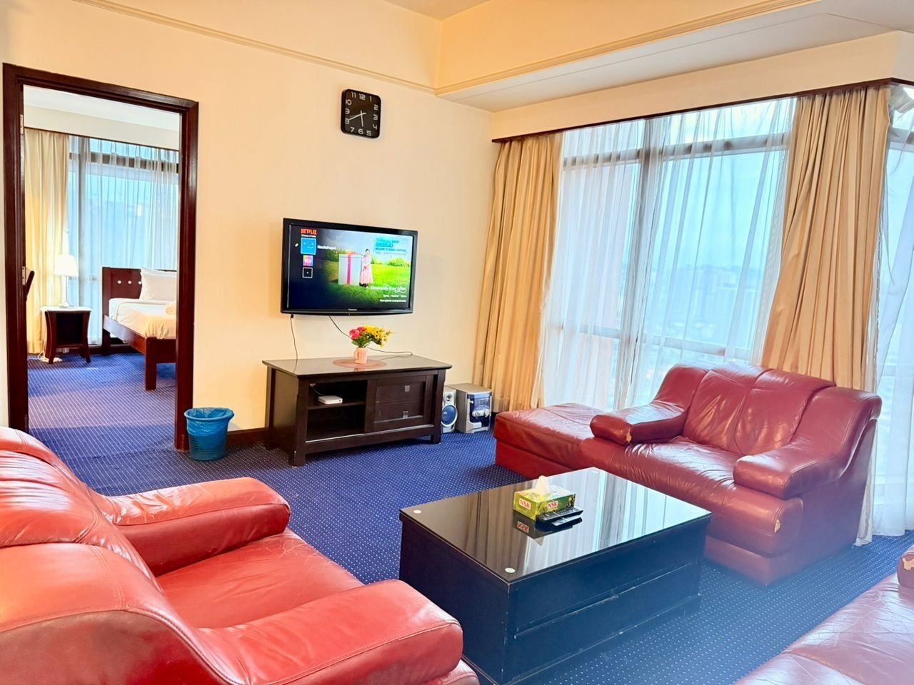A hotel room with a red leather sofa, a flat-screen TV, a small table with a vase, and a window with curtains.