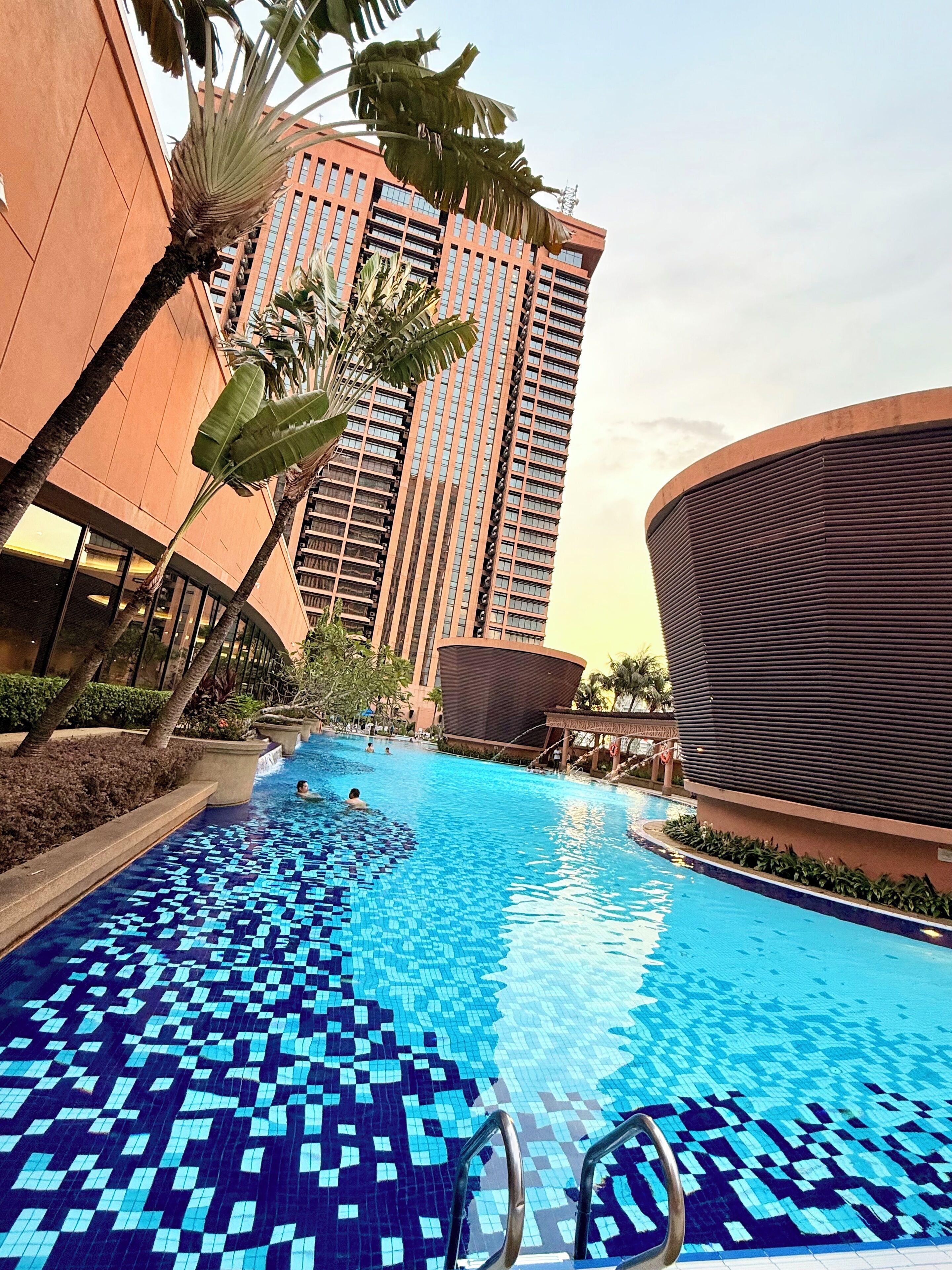 Una piscina con mosaicos azules, rodeada de palmeras y edificios modernos.