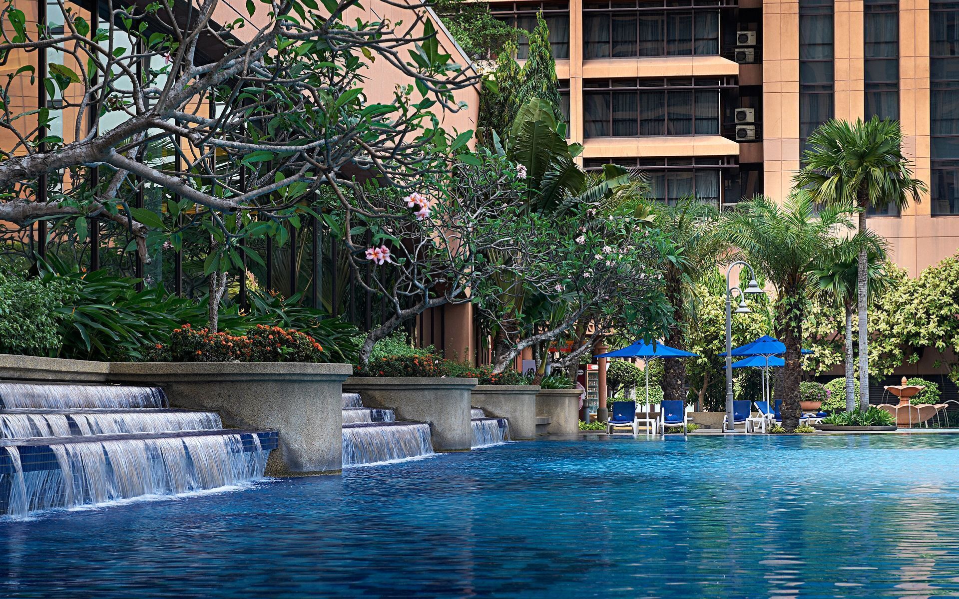 A pool with a waterfall feature, surrounded by lush greenery and a modern building with multiple floors.
