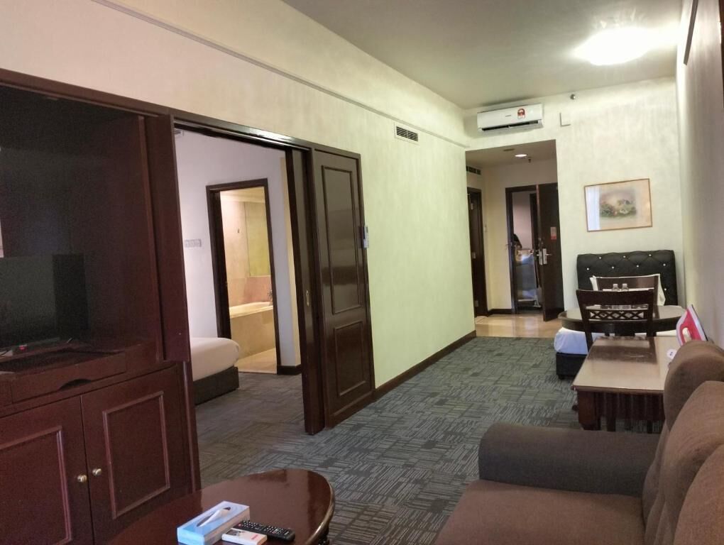 A hotel room with a large wardrobe, a sofa, a coffee table, and a TV. There is a door leading to another room and an air conditioning unit on the wall.