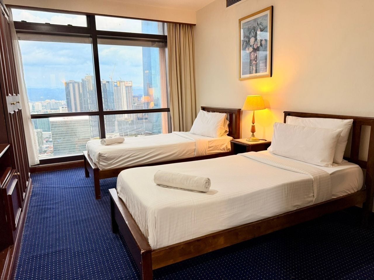 A hotel room with two beds, a large window with a city view, a bedside lamp, and a framed picture on the wall.