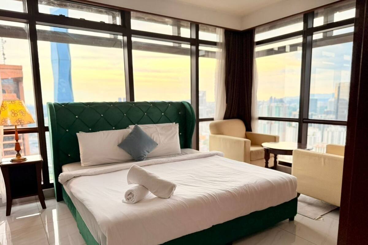A hotel room with a large bed, a green upholstered headboard, a white bedspread, a blue pillow, a small round table, and a chair.