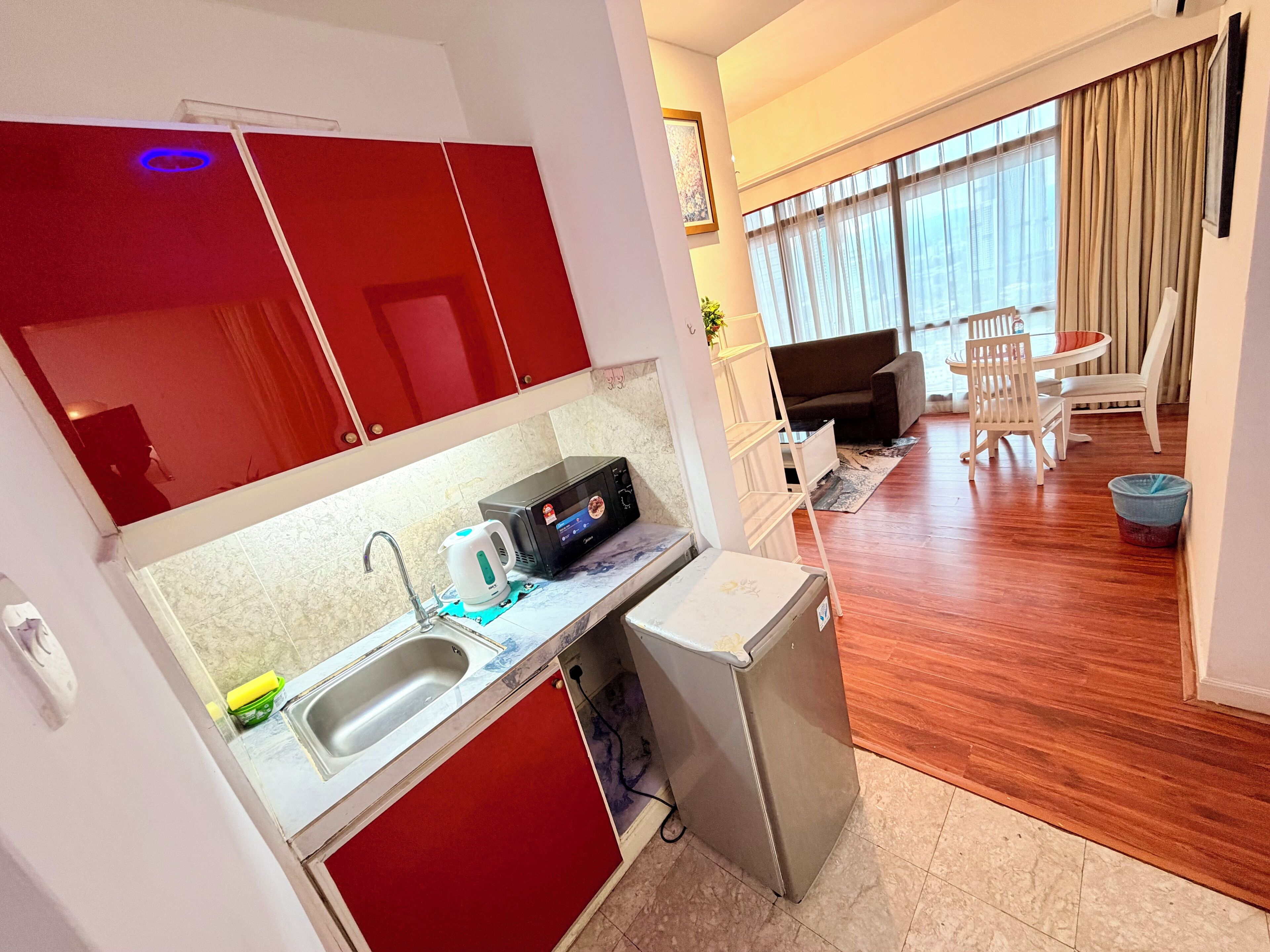 A compact kitchen with a microwave, kettle, and sink. A small refrigerator is next to the sink. The room has a wooden floor and a dining area with a table and chairs.