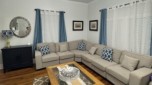 A living room with a sectional sofa, a wooden coffee table, a black cabinet, a mirror, and blue curtains. - Spacious 3-BR Bungalow with Full Amenities Near University Area (Charlotte)