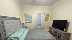 A bedroom with a tufted headboard, a bed with a grey and blue bedspread, a bedside table with a lamp, a television on a cabinet, and a window with curtains. - Spacious 3-BR Bungalow with Full Amenities Near University Area (Charlotte)