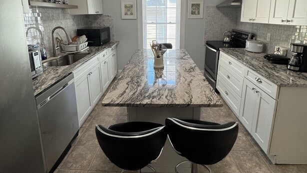 A modern kitchen with white cabinets, a marble island, two black bar stools, and a built-in oven. - Spacious 3-BR Bungalow with Full Amenities Near University Area (Charlotte)