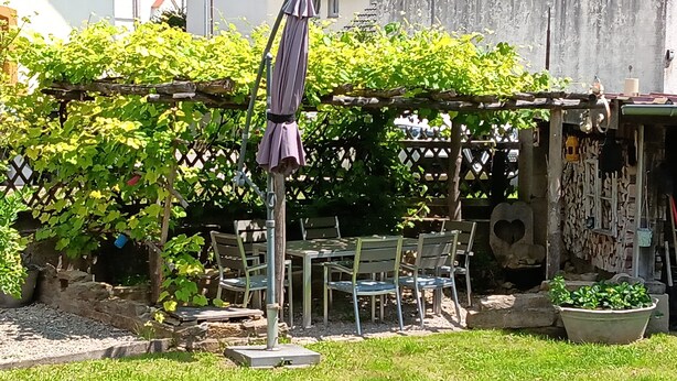 Terassialue, jossa on pöytä ja tuoleja, ja jota ympäröi vihreää kasvillisuutta sekä pergola.