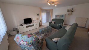 A living room with a sofa, a coffee table, a TV on a white cabinet, and a window with curtains. - Family-friendly vacation home Bornholzweg in Quedlinburg - 157 sqm (Quedlinburg)