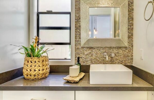 A bathroom with a large mirror, a white sink, a woven basket with a plant, and a soap dispenser.