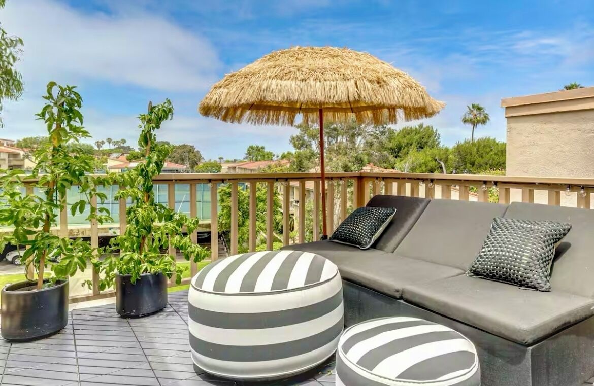 A sunlit outdoor seating area with a thatched umbrella, a cushioned sofa, and striped ottomans.