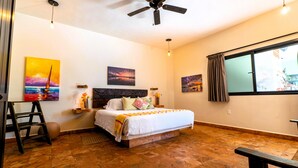 Une chambre à coucher avec un lit, un ventilateur de plafond, des œuvres d’art accrochées aux murs, une fenêtre avec des rideaux et un plancher en bois.