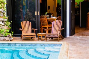 A poolside area with wooden chairs and a dining table, a tiled pool, and a stone wall with plants. - One bedroom suite in Punta Sur (Isla Mujeres)