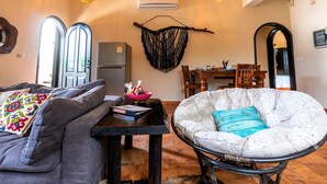 A living room with a grey sofa, a white armchair with a turquoise pillow, a black coffee table, and a cowhide rug. There is a dining area with a wooden table and chairs, and a kitchen with a refrigerator. - 2BR Suite in Punta Sur w/Pool (Isla Mujeres)
