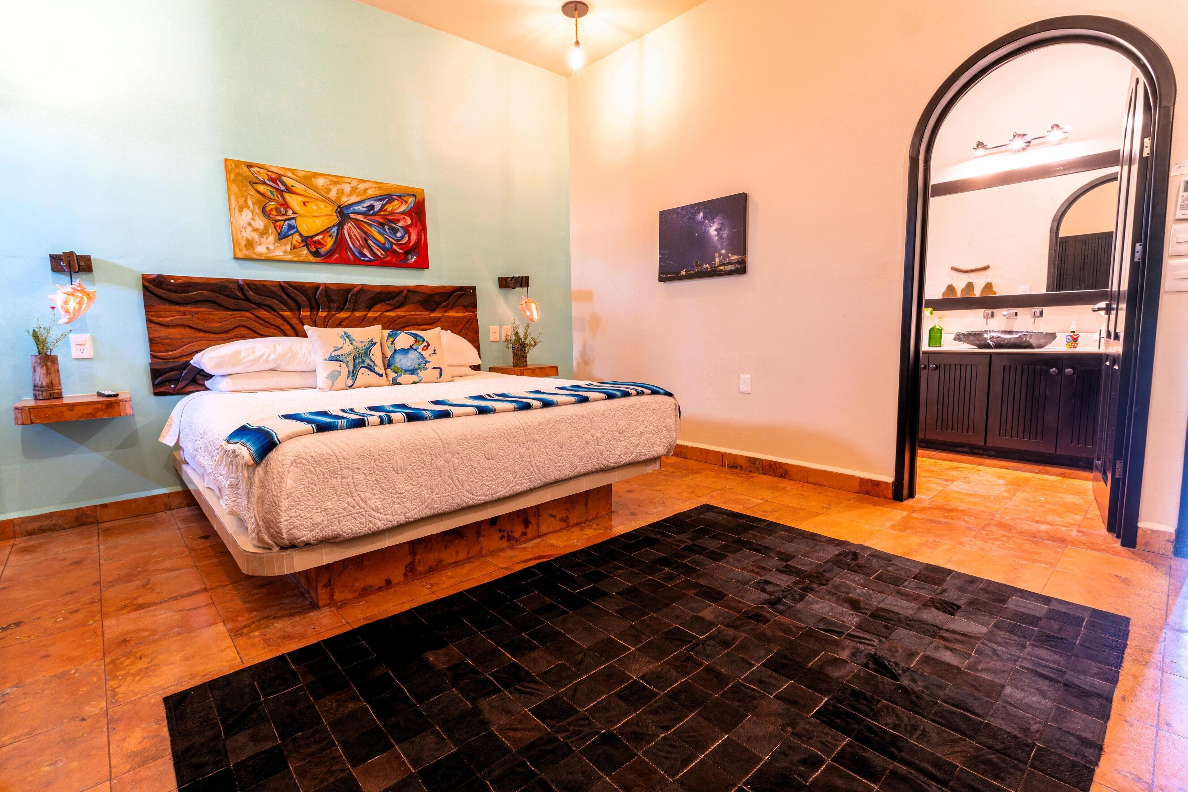 Une chambre à coucher avec un lit, un tapis noir, un tableau, et une salle de bain visible à travers une arche.