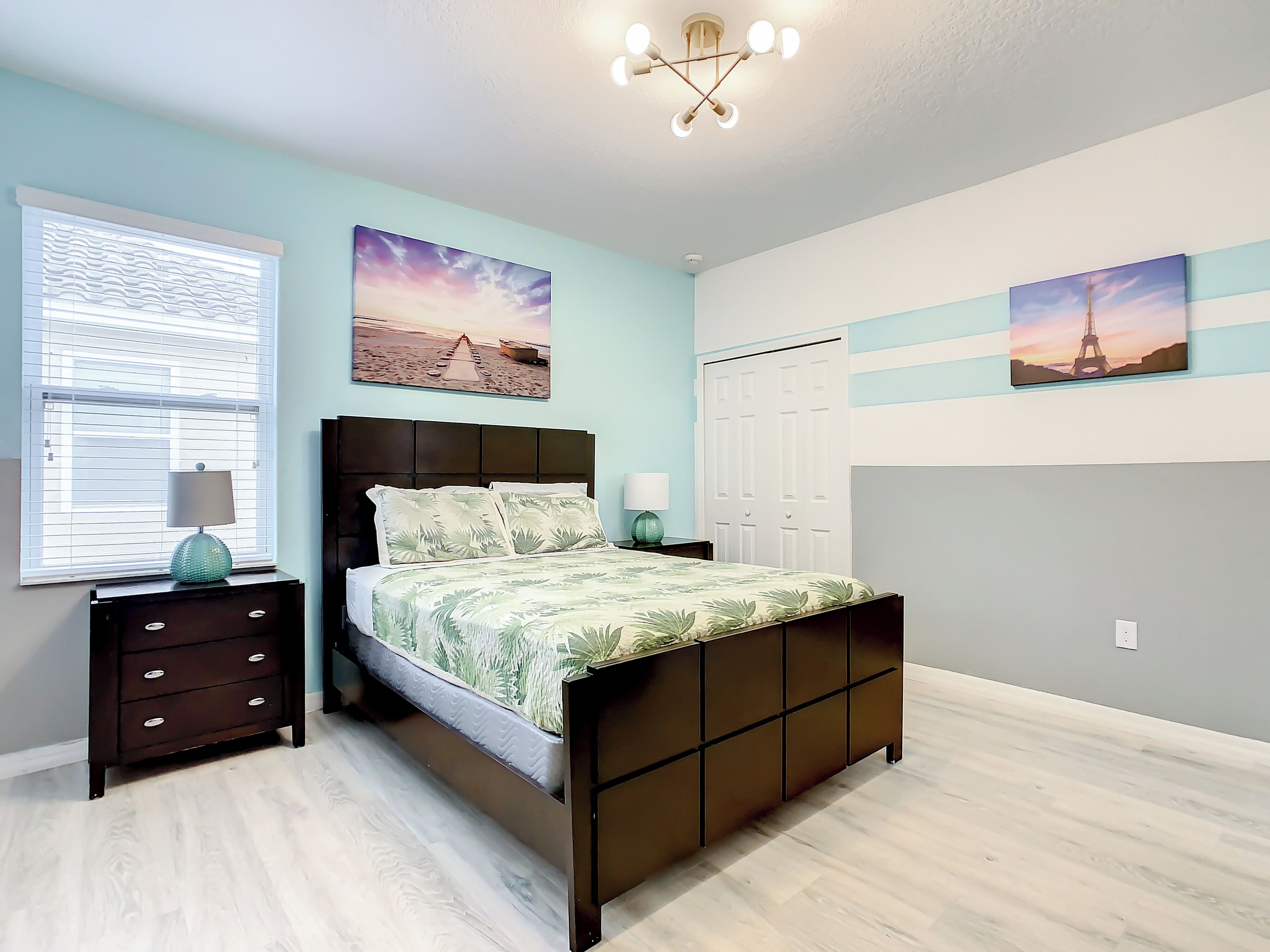 Une chambre à coucher comprenant un lit, des tables de chevet, une fenêtre avec des stores et des œuvres d’art murales.