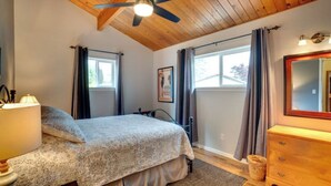 Une chambre à coucher comprenant un lit, une commode, un ventilateur de plafond et deux fenêtres avec des rideaux.