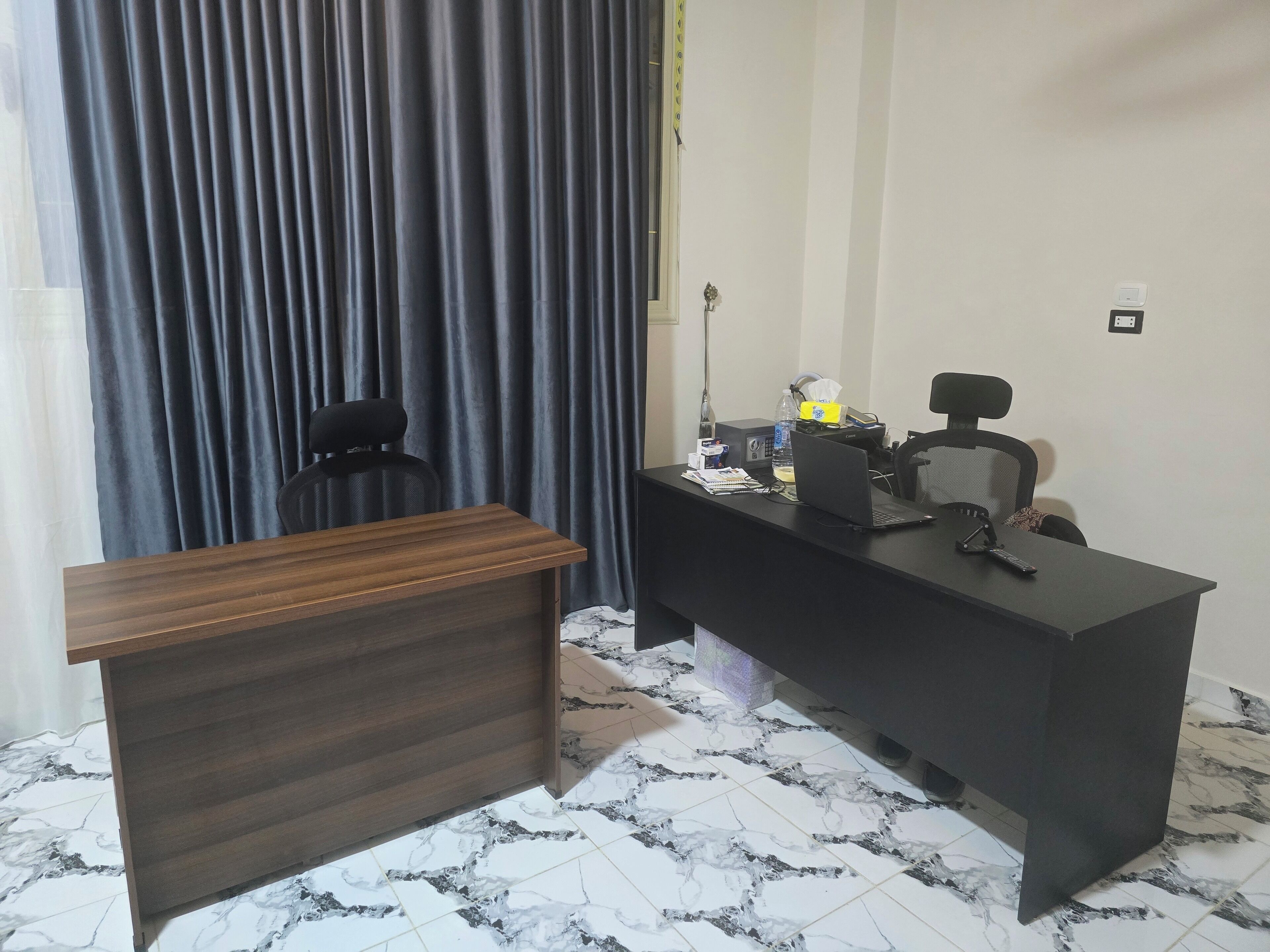 Een moderne kantoorruimte met een houten bureau, een zwarte bureaustoel, en een bureau met een laptop, telefoon en diverse spullen erop.