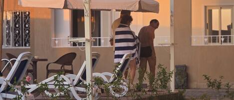 Un espace aménagé au bord de la piscine, avec des chaises longues et des parasols, un bâtiment doté de balcons, et un chemin menant à la piscine.