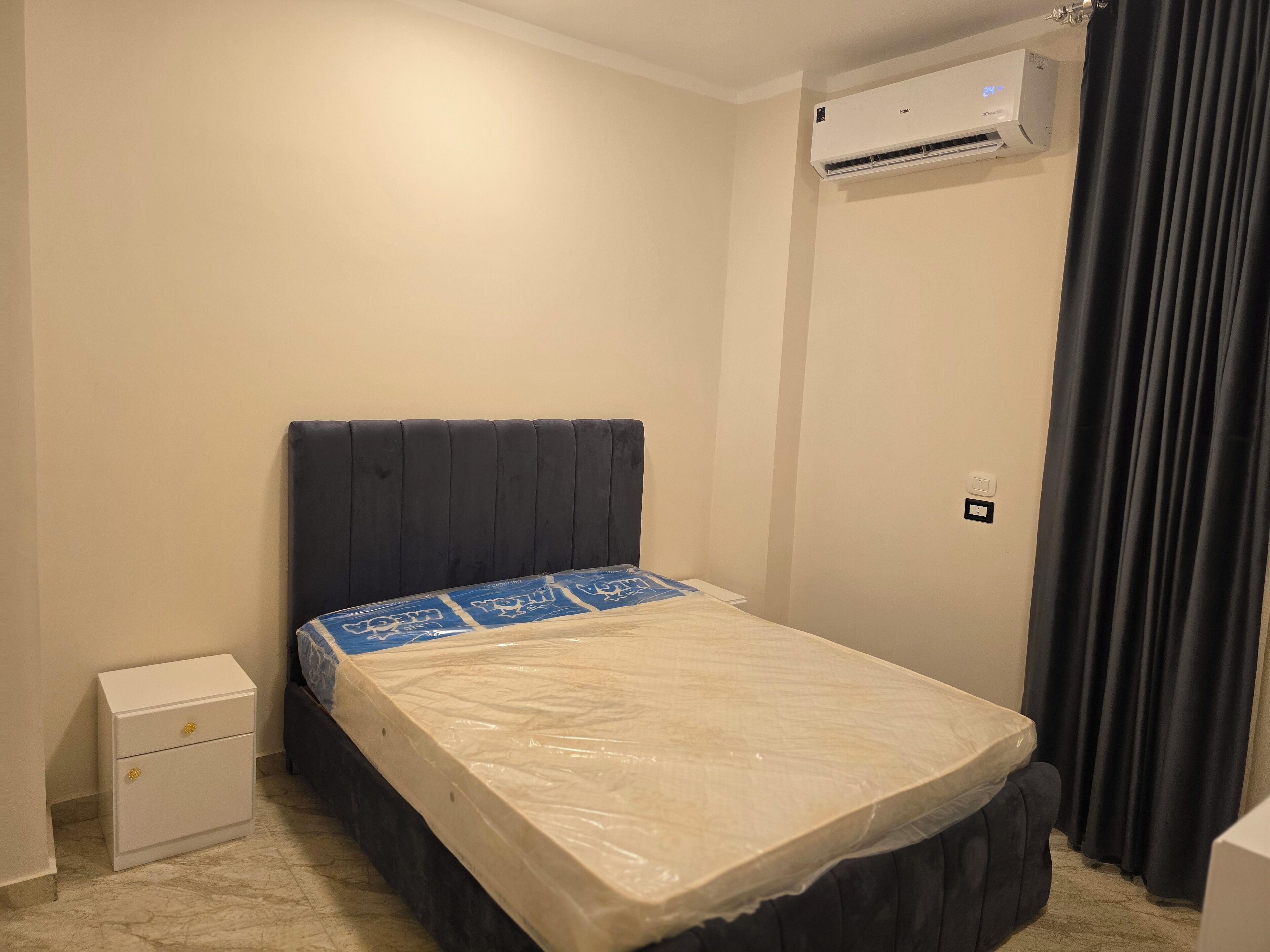 A small, clean room with a bed, a nightstand, an air conditioning unit, and a curtain.