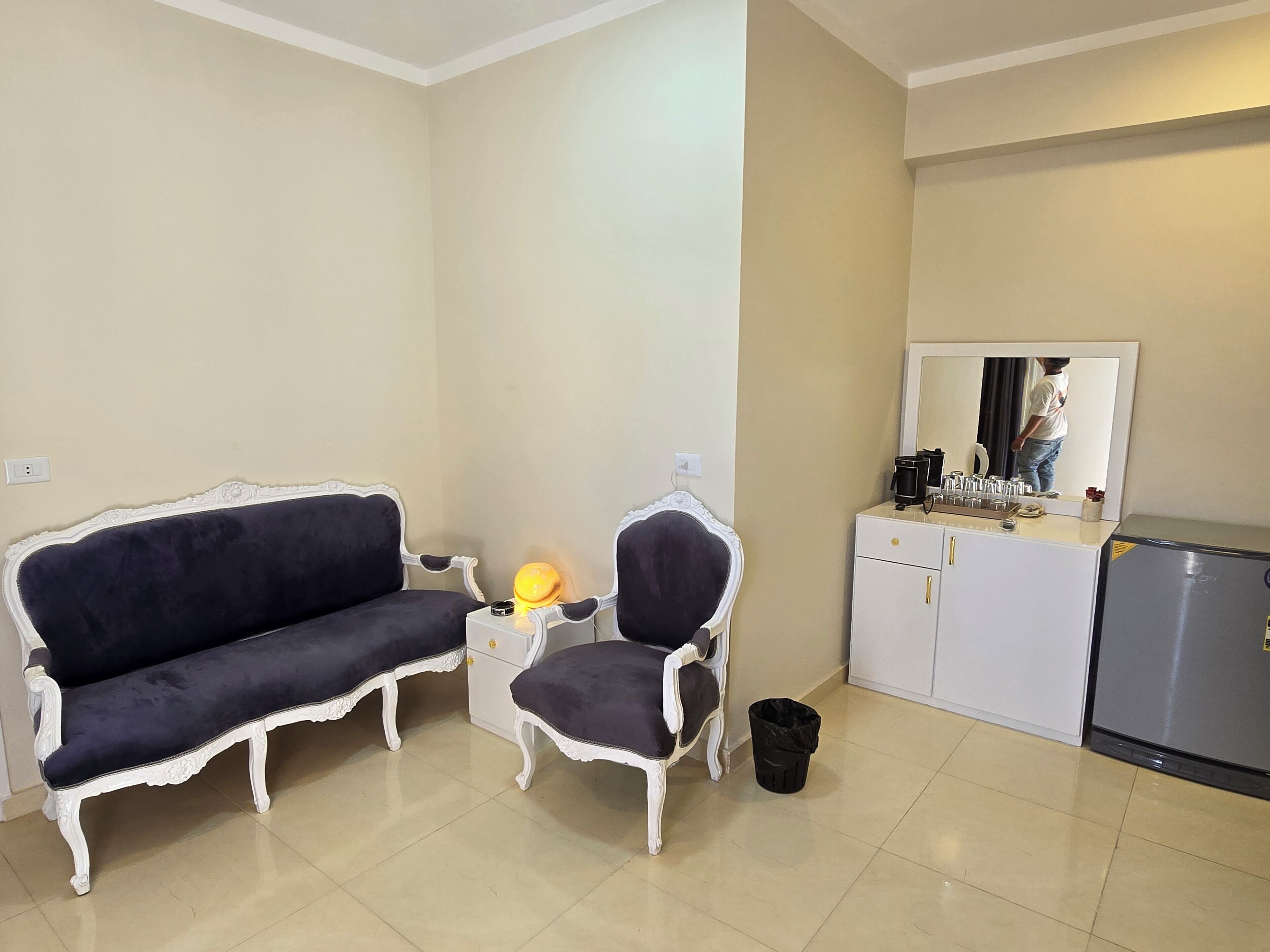 A room with a dark blue sofa, a matching armchair, a small table with a lamp, a white cabinet with a mirror, and a trash bin.